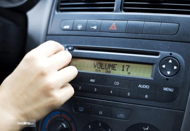 Gambar ini menunjukkan sebuah tangan memutar knob volume pada audio mobil