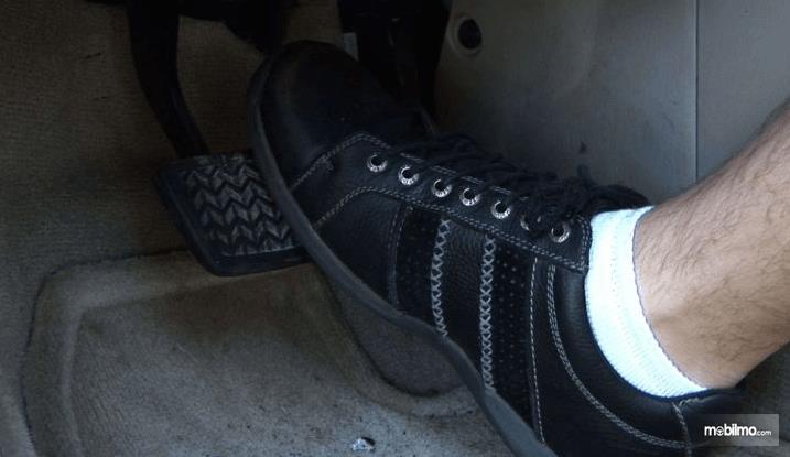 Gambar ini menunjukkan sebuah kaki dengan sepatu hitam diletakkan di atas pedal