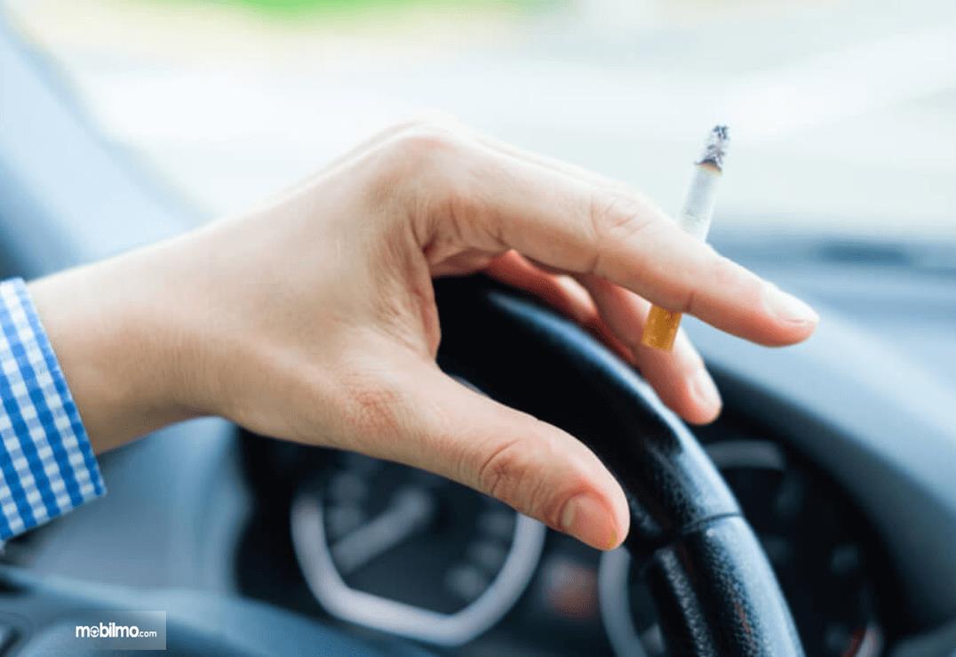 Gambar ini menunjukkan sebuah tangan memegang rokok dan kemudi mobil